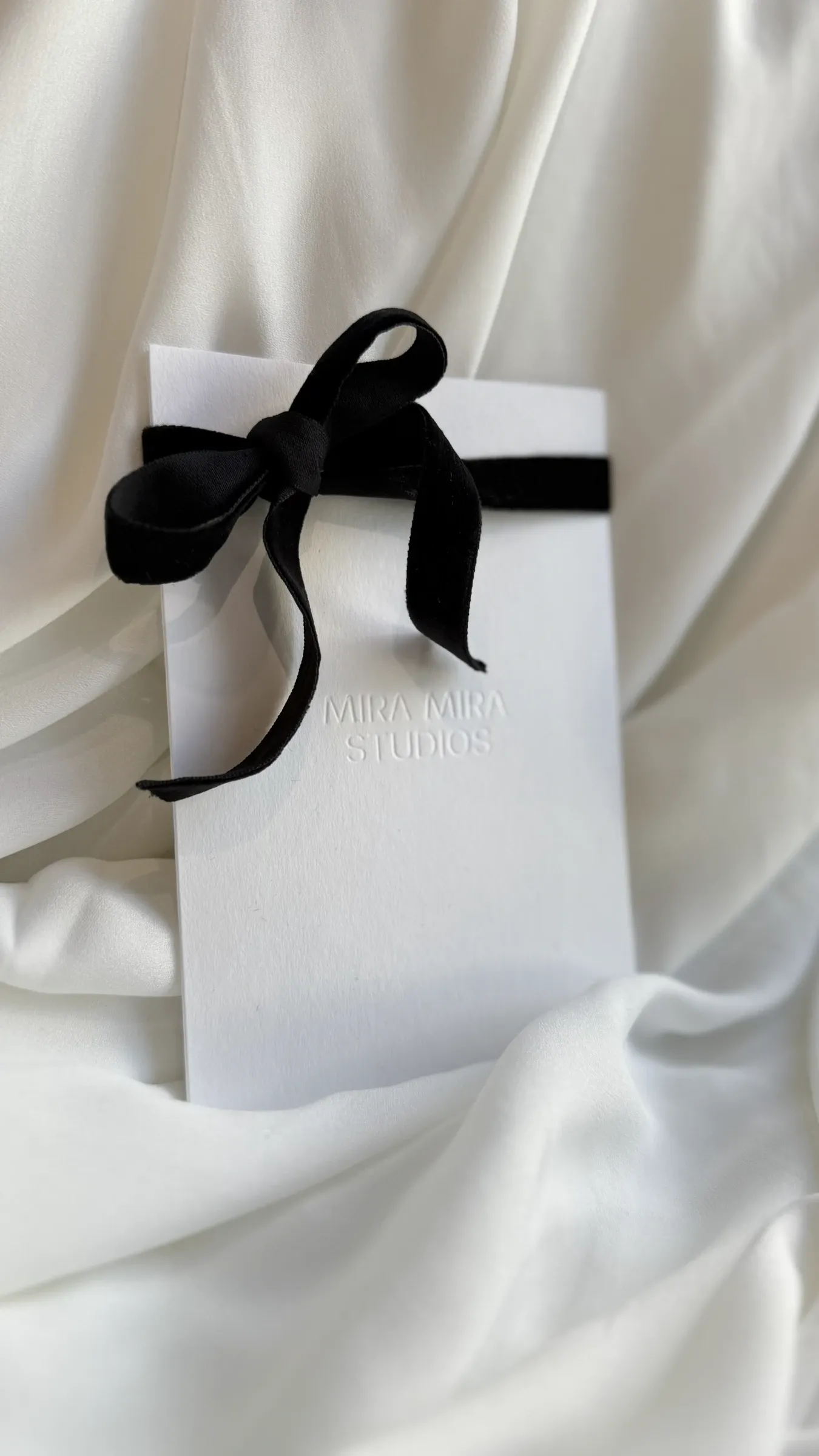 A Mira Mira Studios gift card tied with a black velvet ribbon, resting on white silk